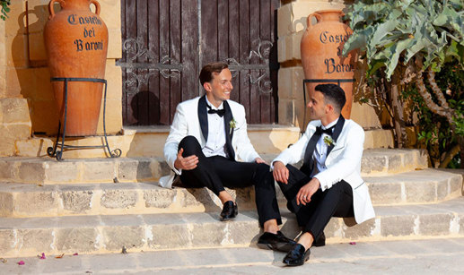 two grooms wearing white suit jackets and bow ties sitting on a set of stone steps in front of a large wooden door. There are two large terracotta urns reading 'Castello dei Baroni' either side