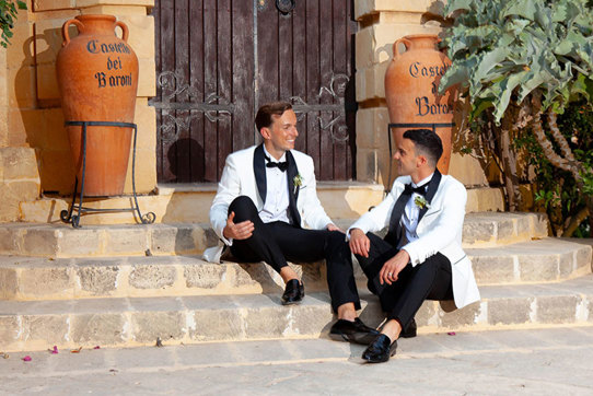 two grooms wearing white suit jackets and bow ties sitting on a set of stone steps in front of a large wooden door. There are two large terracotta urns reading 'Castello dei Baroni' either side