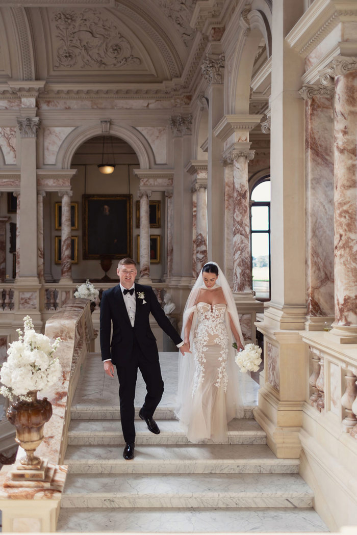 A bride and groom walking down a marble staircase at Gosford House. Bride is wearing a Berta Privee dress from Opus Couture and groom is wearing a black tuxedo from Forbes Tailoring.