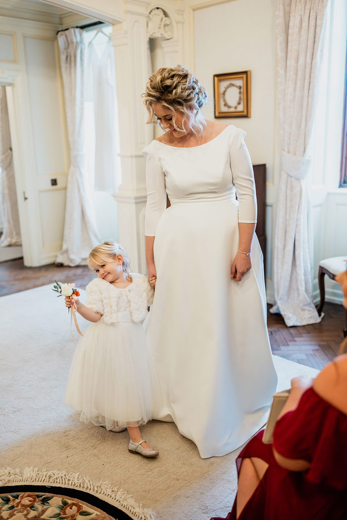 bride in white three-quarter-sleeve wedding dress with her hair up holds hand of young flower girl in little white dress 