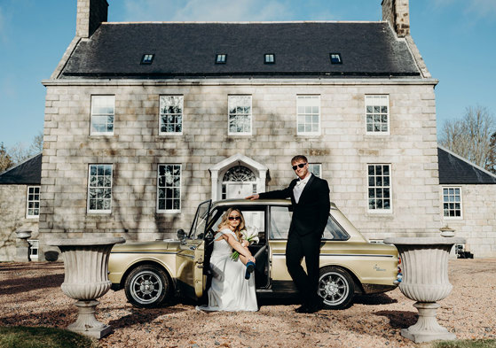 a bride and groom wearing sunglasses posing by a vintage yellow car outside Elrick House in Aberdeen