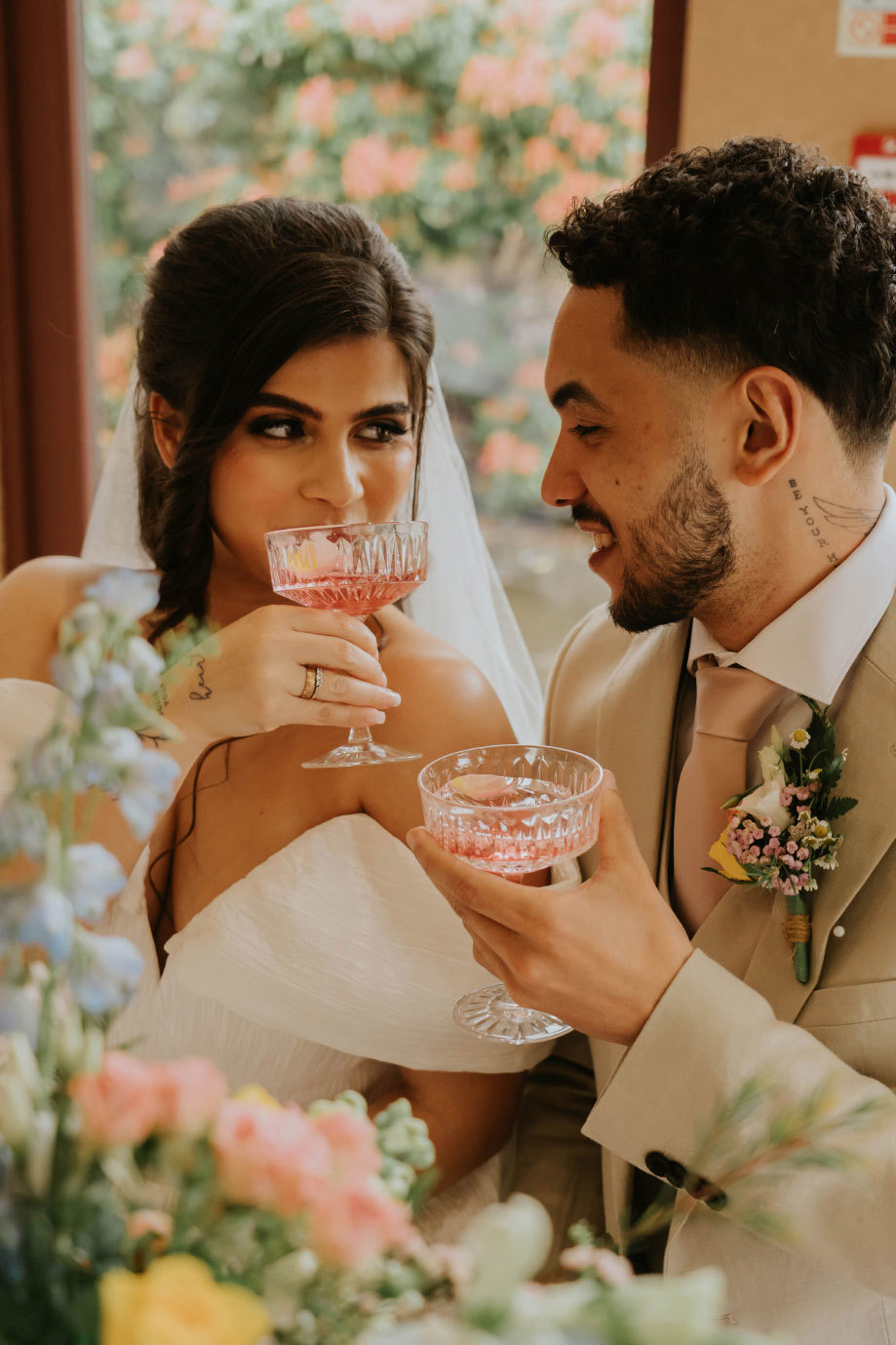 A bride and groom drinking rose champagne from coupe glasses while looking at each other