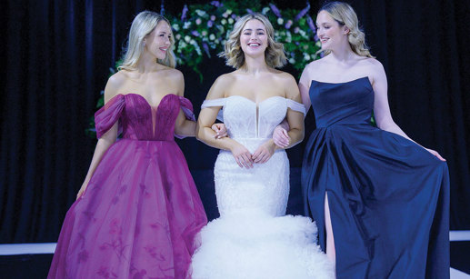 Three models smiling and walking arm in arm on the catwalk at The Scottish Wedding Show. Middle model is wearing an off-shoulder ivory gown with tulle tiered fishtail skirt. Model on right is wearing a navy bridesmaid dress. Model on left is wearing a purple off shoulder dress with floral pattern