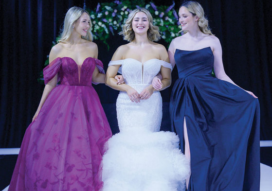 Three models smiling and walking arm in arm on the catwalk at The Scottish Wedding Show. Middle model is wearing an off-shoulder ivory gown with tulle tiered fishtail skirt. Model on right is wearing a navy bridesmaid dress. Model on left is wearing a purple off shoulder dress with floral pattern