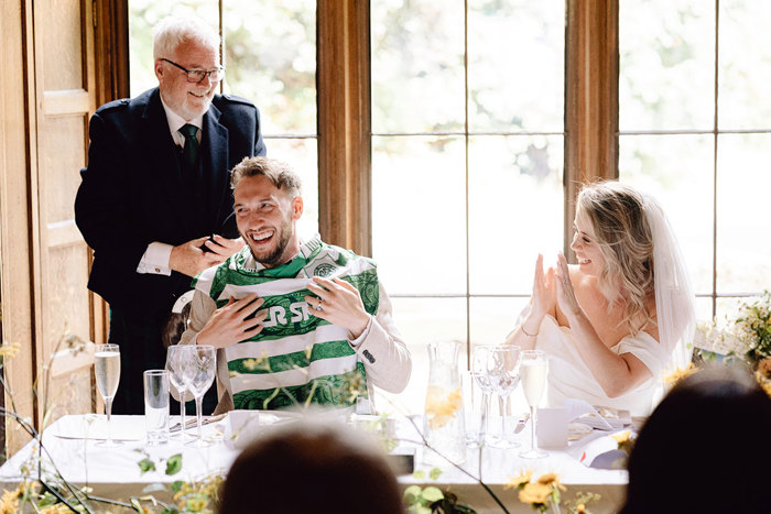 bride and groom laugh as groom is presented with football top during wedding dinner