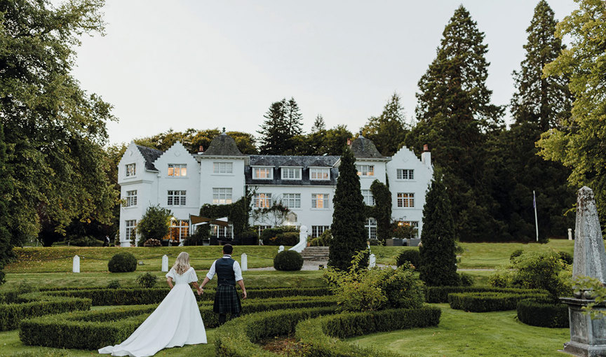 A bride and groom in formal attire walking in a garden in front of the white exterior of Achnagairn Castle