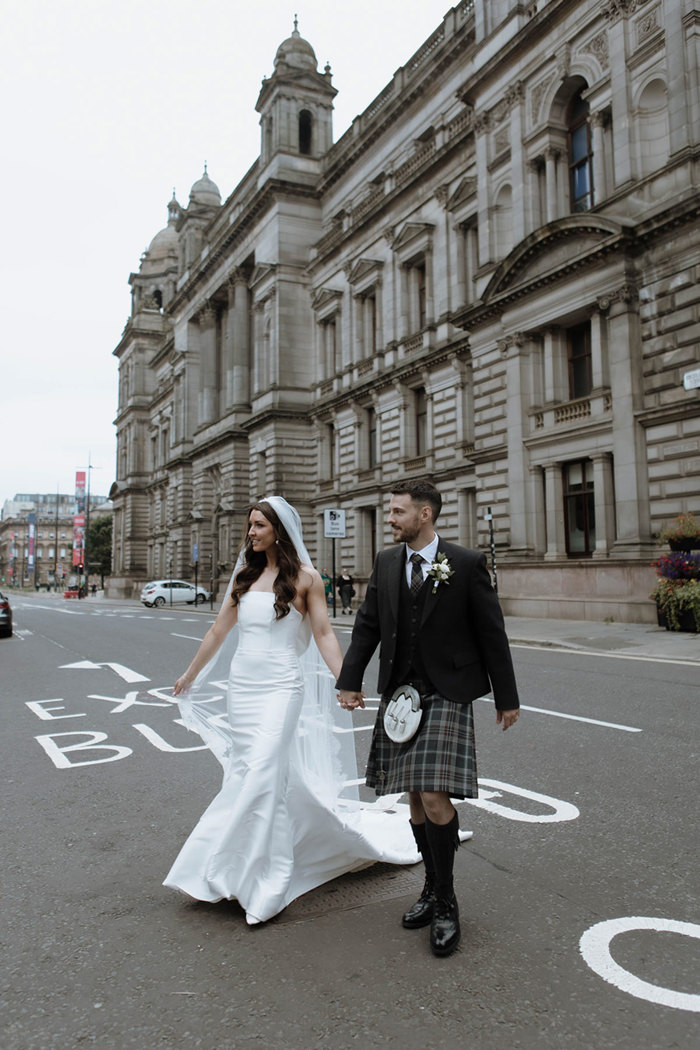 A bride in a fitted dress and a groom in a dark kilt cross a road holding hands