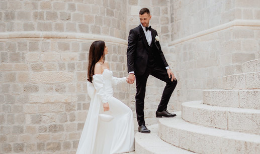 A couple in formal attire poses on the stone steps of Dubrovnik; the woman wears a white dress, and the man dons a black tuxedo.