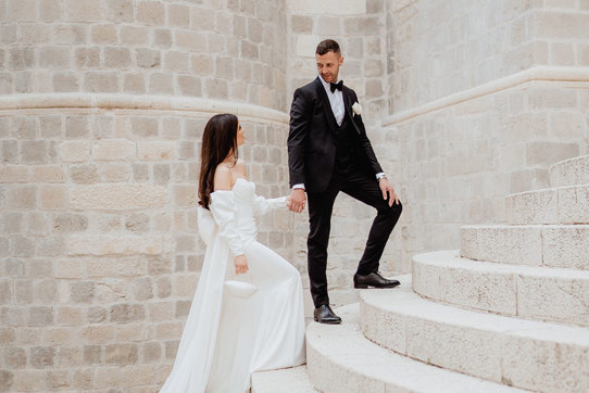 A couple in formal attire poses on the stone steps of Dubrovnik; the woman wears a white dress, and the man dons a black tuxedo.