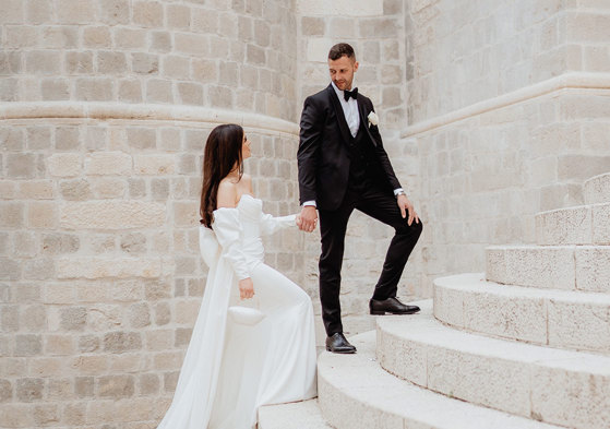 A couple in formal attire poses on the stone steps of Dubrovnik; the woman wears a white dress, and the man dons a black tuxedo.