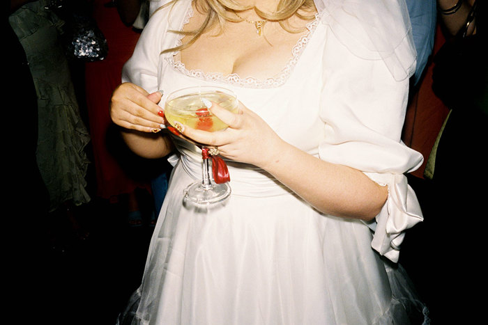 shoulder-down view of bride in square-neck wedding dress holding a coupe glass of champagne with a cocktail cherry in it