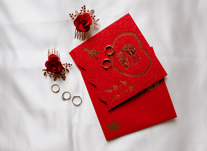 embossed red and gold wedding invite on a white fabric background with small red rose hair combs and engagement and wedding rings