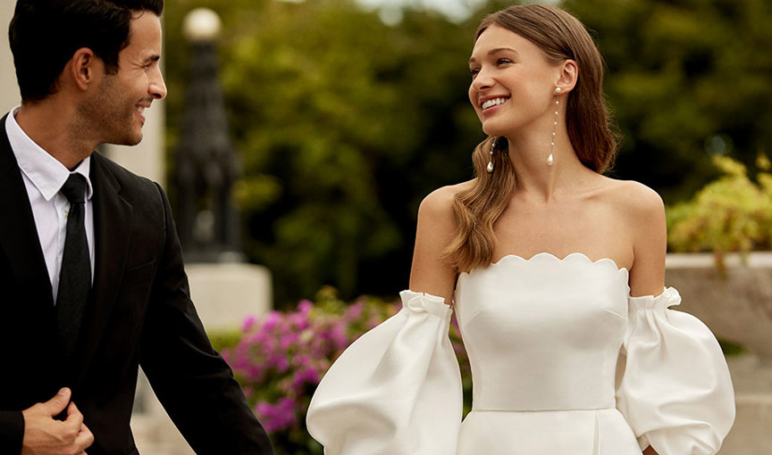 a person wearing a smart black suit holding hands and walking with a person in an ivory Rosa Clara dress with removable puffed sleeves