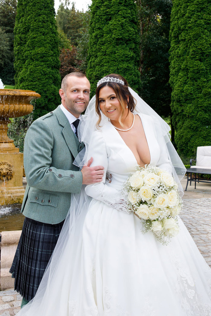 a bride in a V-neck white dress and groom in a kilt posing for a photo.