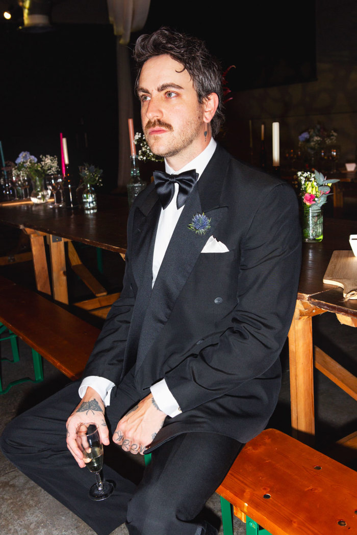 a man with a moustache wearing a black suit and bow tie sitting on a bench and holding a glass of champagne