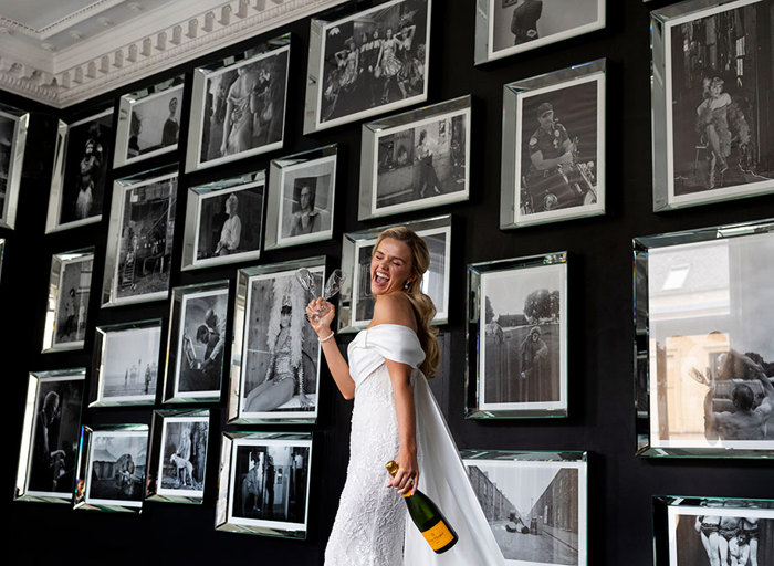 a model wearing a wedding dress and holding champagne bottle and glasses in front of a wall of black and white framed photographs