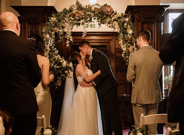 A bride and groom kiss underneath a flower arch