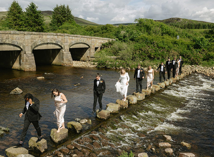 a line of people wearing smart black dinner suits and bow ties and long cream dresses walking across stepping stones across a flowing  river. There is an arched stone bridge, trees and hills in background