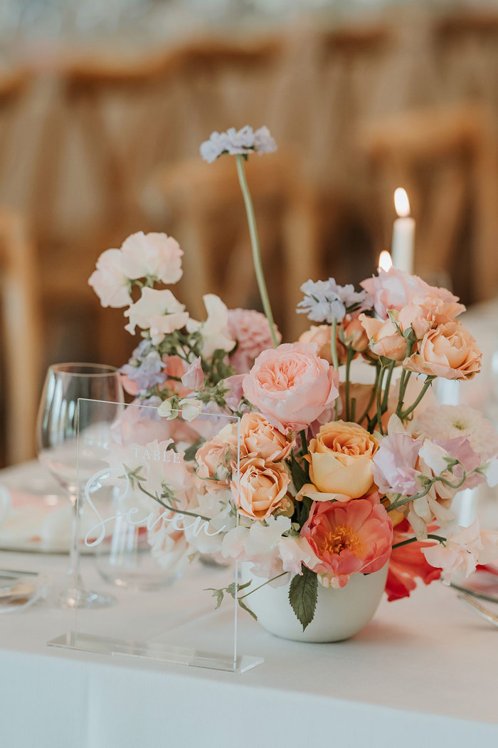 Pink, orange and purple flowers in a white vase on a table with a white table cloth and drinking glasses on it