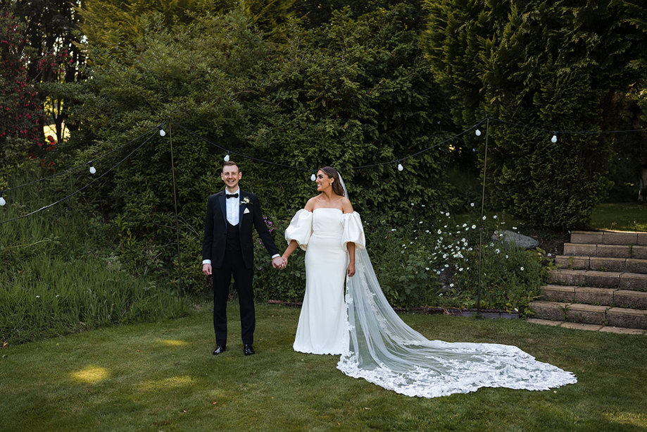 Bride looks at her groom smiling and holding hands in garden