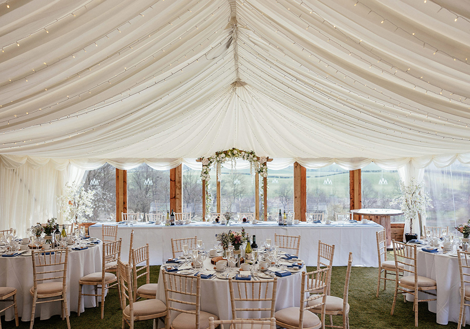 Inside a white marquee with a long top table and round tables all with white table cloths, white chiavari chairs and greenery centrpieces