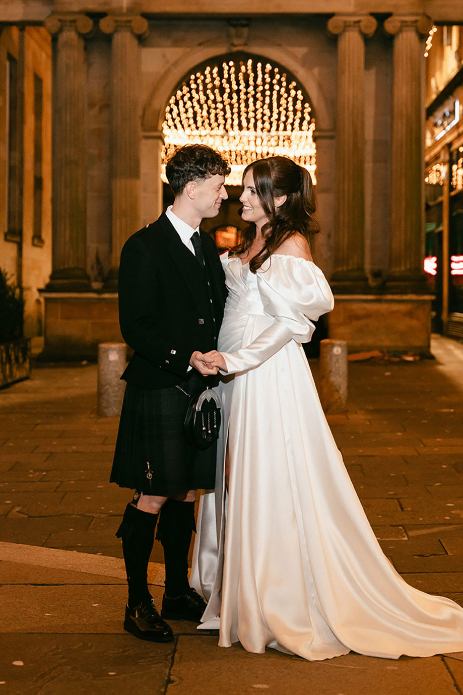 brunette bride in high leg slit wedding dress with long puff sleeves and groom in full dark tartan kilt outfit stand close facing and smiling at each other outdoors with a canopy of twinkling lights in the night sky above them