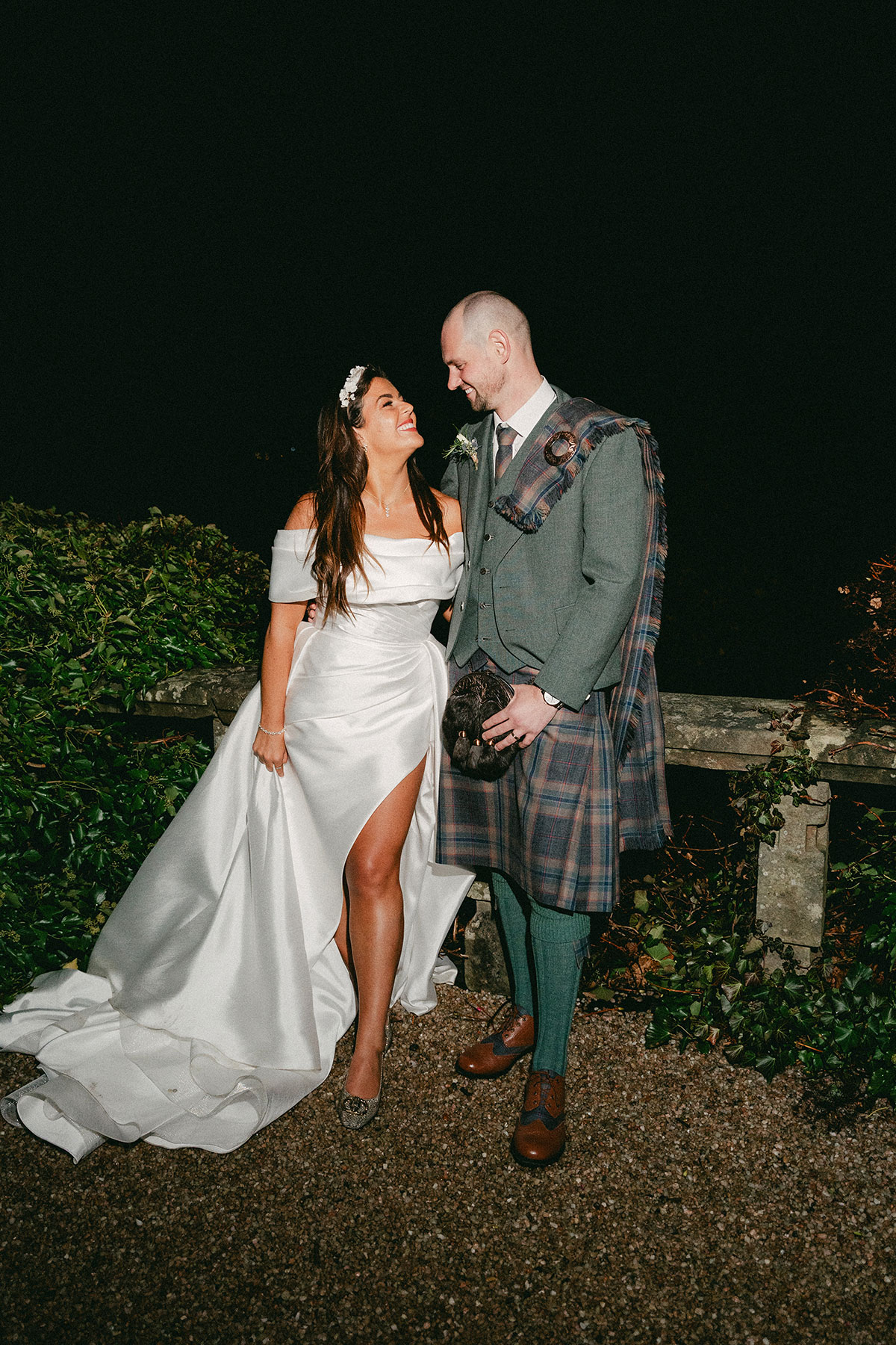 bride with floral headband and off the shoulder wedding dress with a leg slit stands outdoors smiling up at groom in green, blue and purple tartan kilt outfit with green jacket