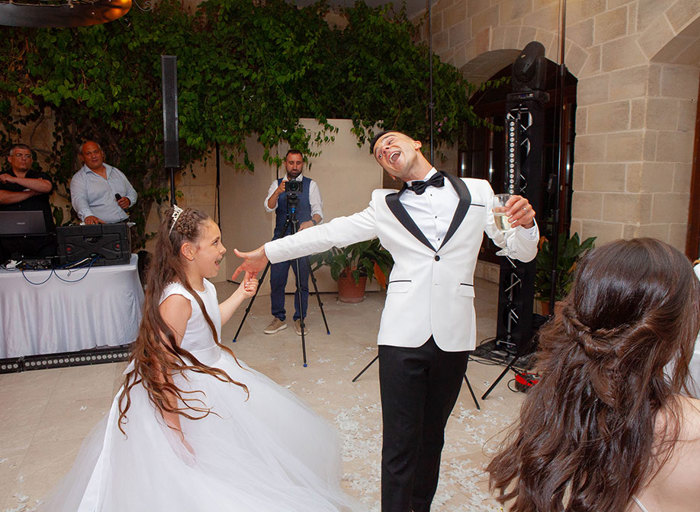 a man wearing a white tuxedo jacket and bow tie dancing with a flower girl wearing a white dress. There is a DJ and sound desk in the background