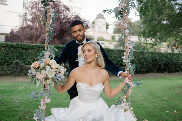 A bride in a sweetheart neckline ballgown dress sits on a swing with flowers and vines entwined in the ropes as she is being pushed by a man in a suit