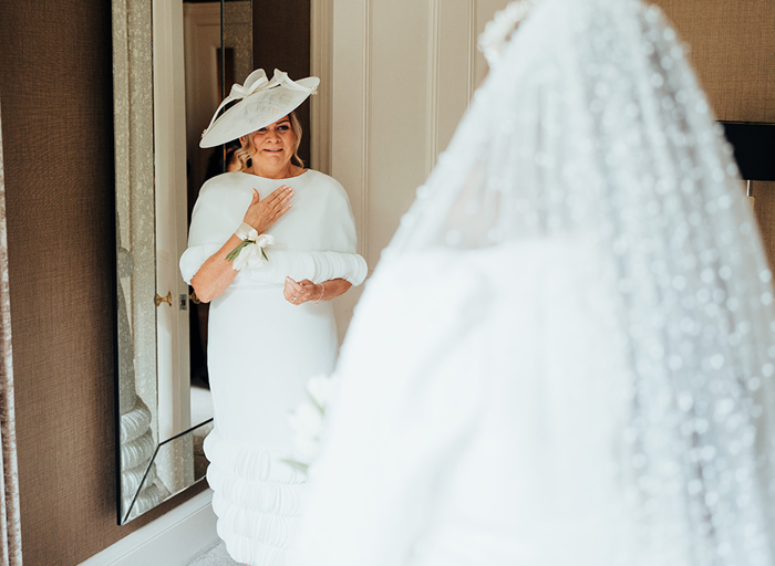  A first look between a bride and her mother, the bride has her back to the camera and the mother of the bride is wearing a white dress and hat and looks emotional