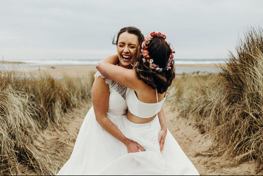 two brides embracing joyfully in a field of tall grass with sand on the ground, one wearing a flower crown