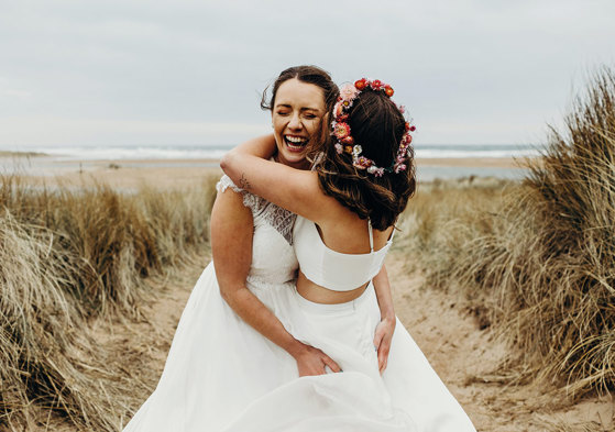 two brides embracing joyfully in a field of tall grass with sand on the ground, one wearing a flower crown