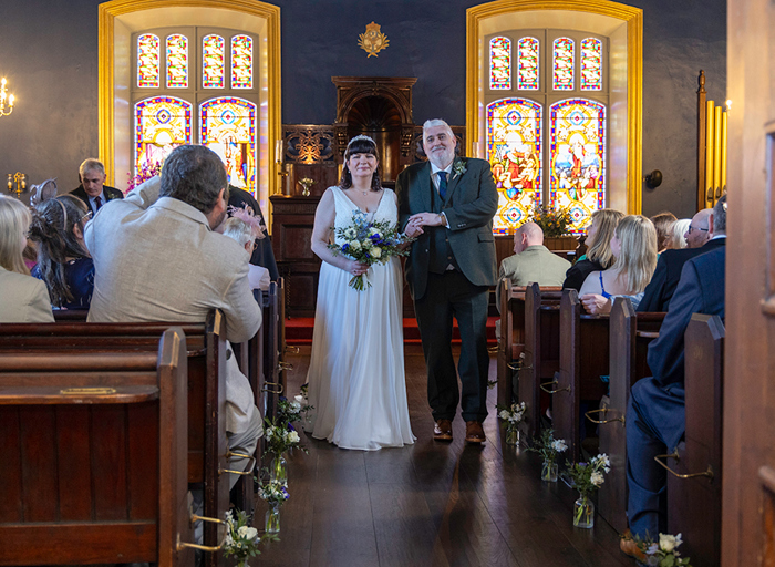 a happy bride and groom walk up the aisle in a church with brightly coloured stained glass windows behind them