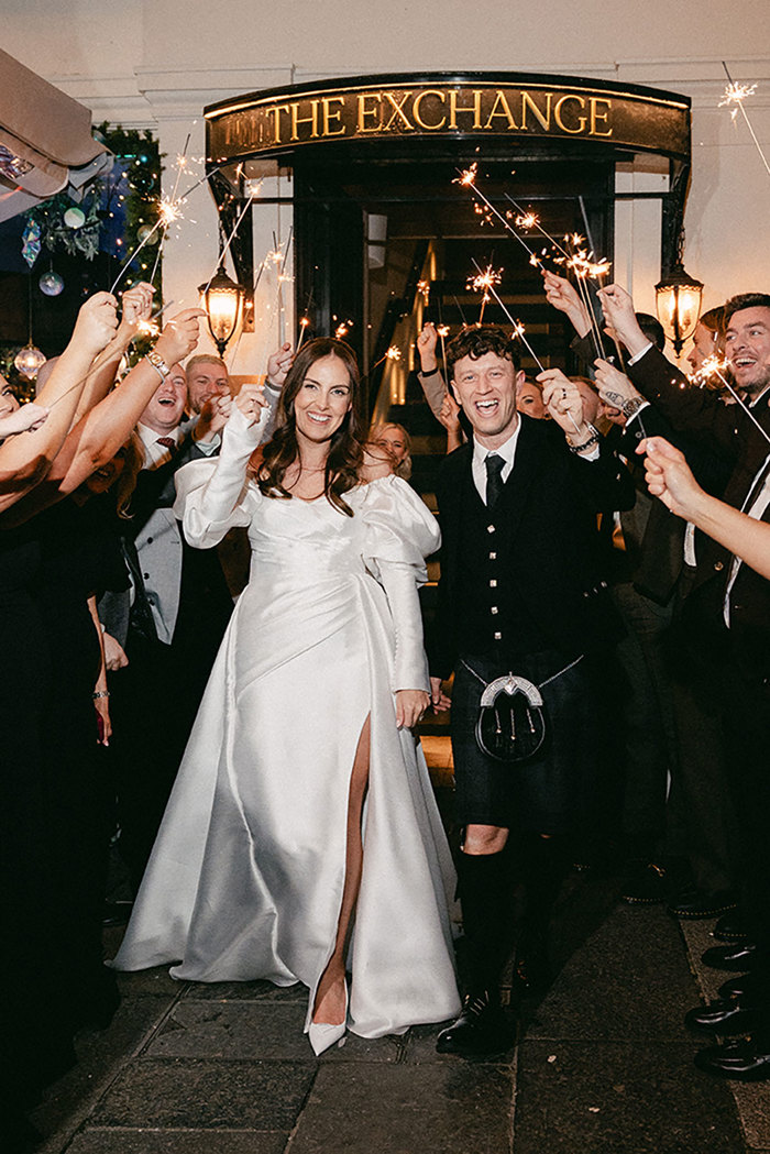 brunette bride in high leg slit wedding dress with long puff sleeves and groom in full dark tartan kilt outfit walk hand in hand through a tunnel of people cheering and holding sparklers up in the air in front of a large sign saying 'The Exchange'