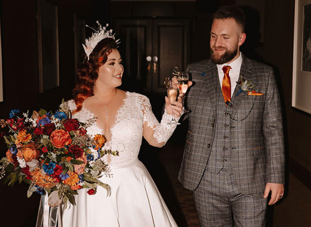 a bride in a white dress holding a large bouquet and groom in a grey suit with an orange tie clink glasses of champagne