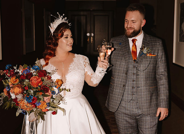 a bride in a white dress holding a large bouquet and groom in a grey suit with an orange tie clink glasses of champagne