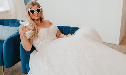 bride sitting in wedding gown on blue couch with pearl sunglasses and a coupe of prosecco