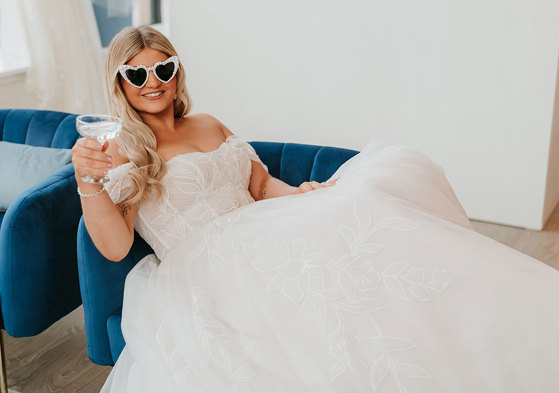 bride sitting in wedding gown on blue couch with pearl sunglasses and a coupe of prosecco