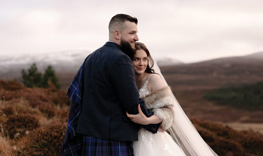 a bride wearing a faux fur wrap, veil and ivory dress snuggles into a groom wearing a blue kilt and tweed jacket. They are standing in an autumnal landscape surrounded by brown bracken and there is snow on distant hills