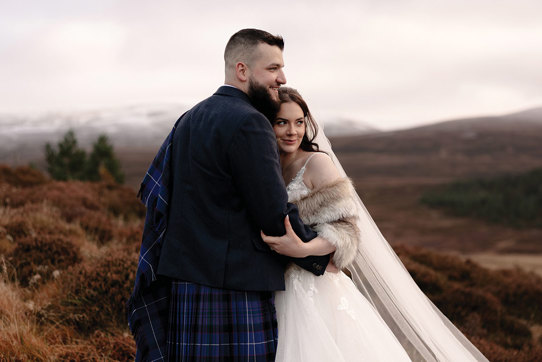 a bride wearing a faux fur wrap, veil and ivory dress snuggles into a groom wearing a blue kilt and tweed jacket. They are standing in an autumnal landscape surrounded by brown bracken and there is snow on distant hills