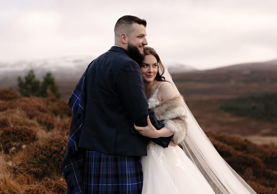 a bride wearing a faux fur wrap, veil and ivory dress snuggles into a groom wearing a blue kilt and tweed jacket. They are standing in an autumnal landscape surrounded by brown bracken and there is snow on distant hills