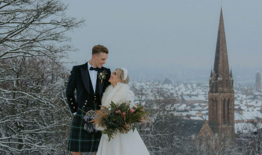 A bride wearing a white dress and white fur wrap standing with a groom wearing a dark kilt in a park in the snow