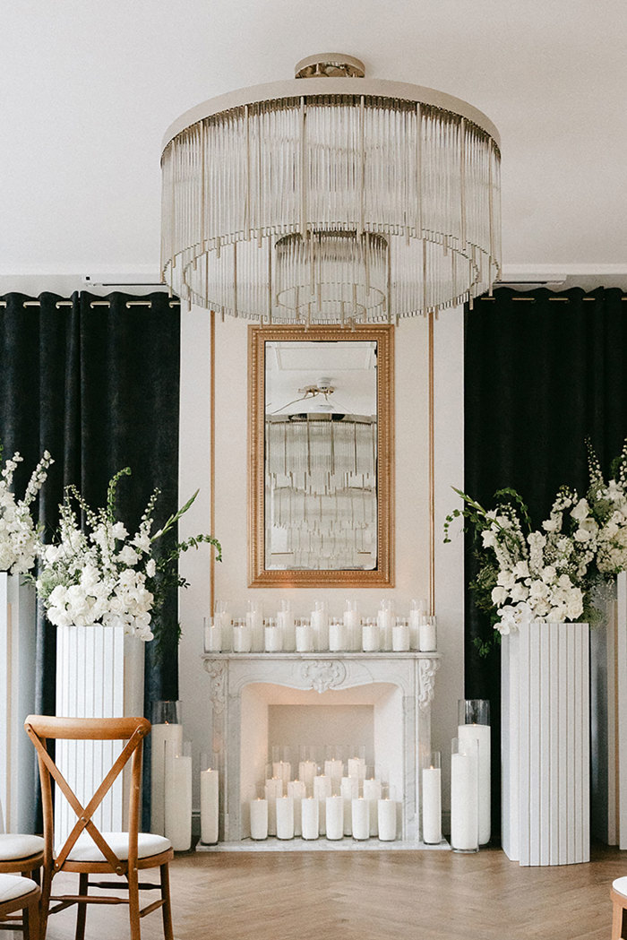 a detail shot of the top of the aisle complete with candle-covered fireplace and mantel, a large gold mirror above, a crystal chandelier, and white podiums of white flowers
