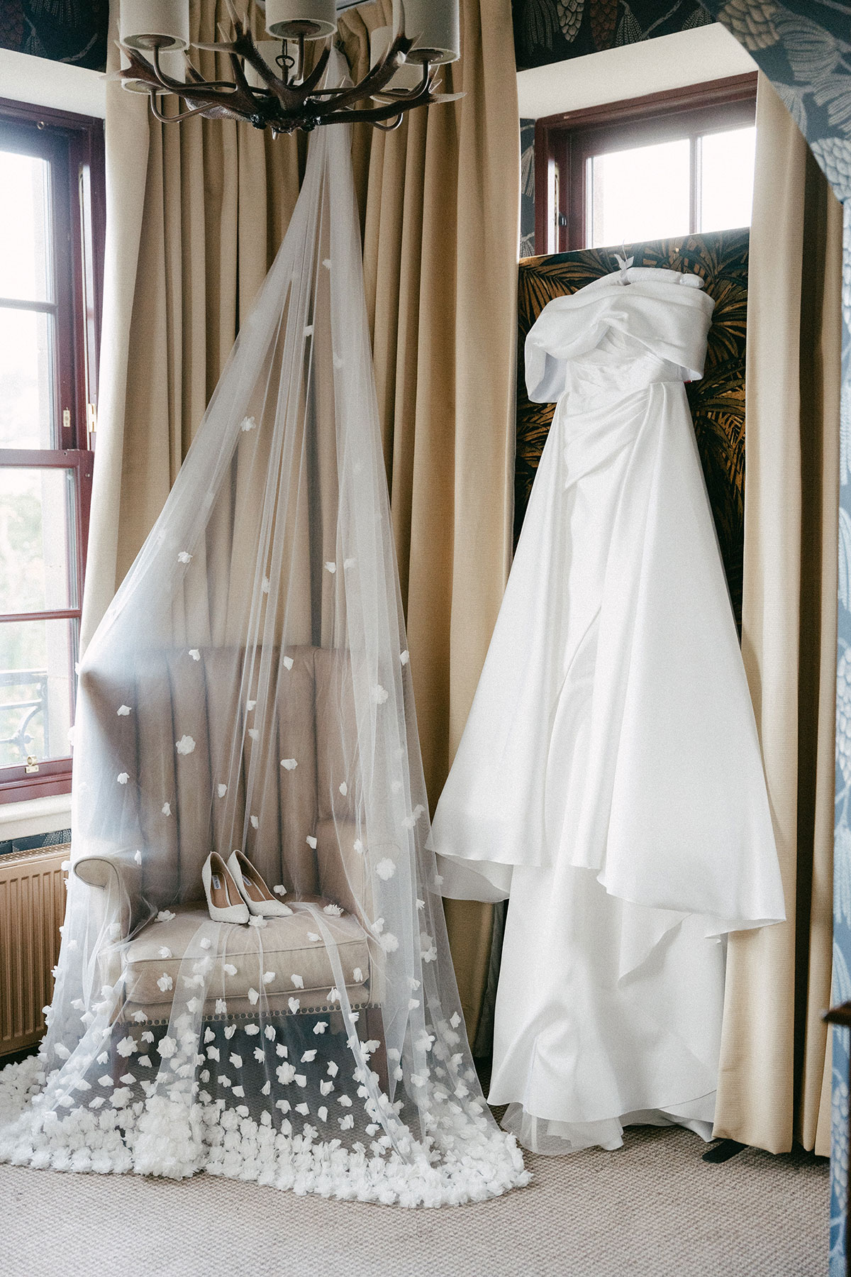 ballgown off the shoulder wedding dress hangs off window blind and long veil hangs off the chandelier and drapes over a lounge chair with a pair of white heels on top