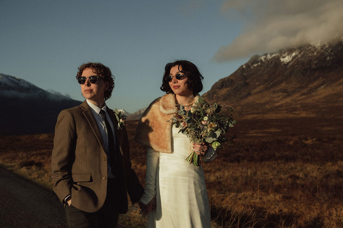 a couple, one in a suit and tie, the other in a white dress and fur shawl holding a bouquet of flowers, both wear sunglasses and looking away from the camera while stood outdoors in the mountains