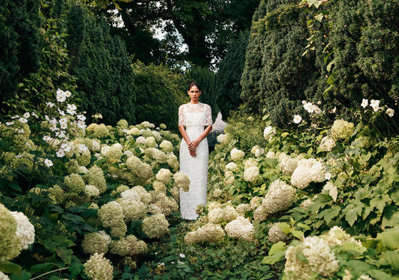 woman in lace wedding dress with lace cape over her shoulders stands in the centre of a green and white flower field with tall hedges around it