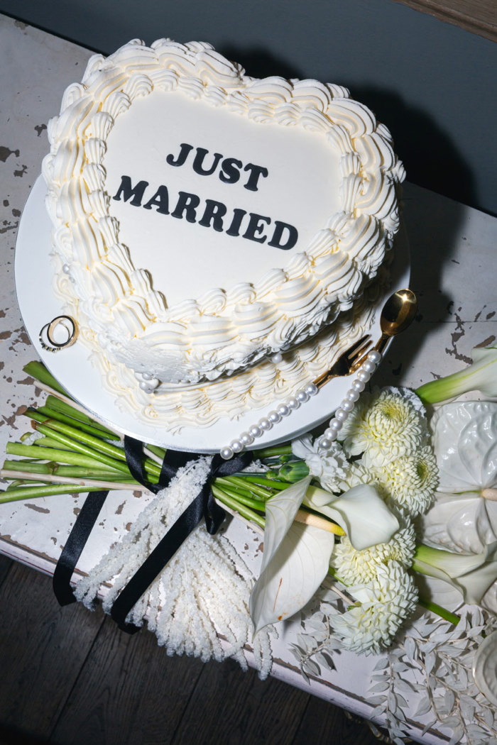 A heart-shaped white lambeth cake with 'Just Married' written on the top in black icing with a pearl embellished spoon and fork, a bouquet of white flowers and two rings sitting next to it