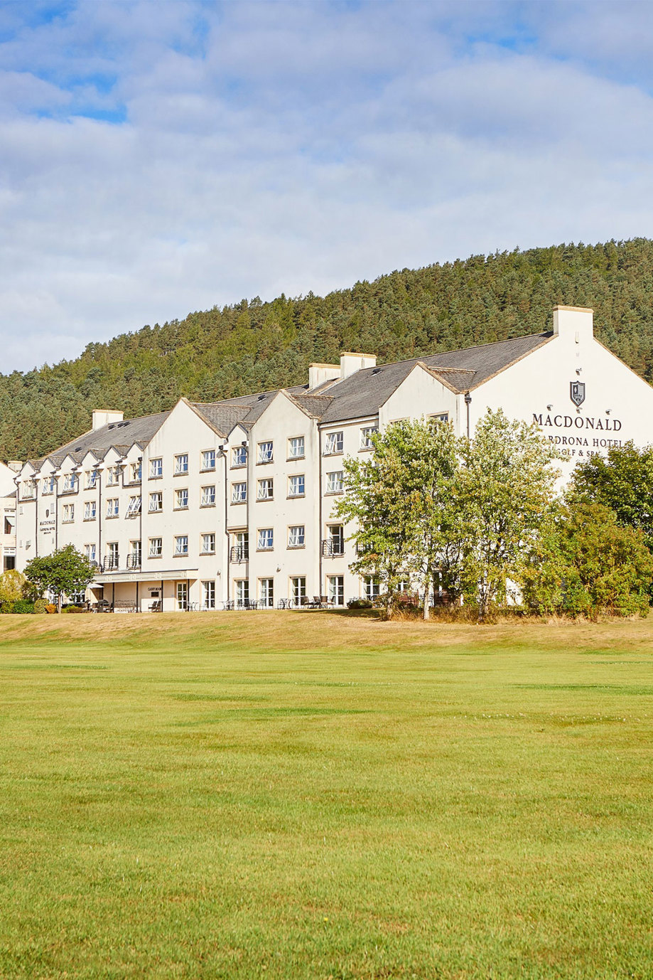 A large white four-storey building with a grey roof with at the bottom of a hill which is covered in trees 