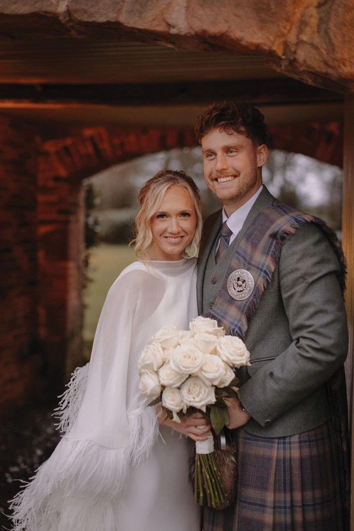 blonde bride in feathery cape and white wedding dress holds bouquet of white roses and stands beside groom in green jacket and red, blue, green kilt with matching fly plaid 