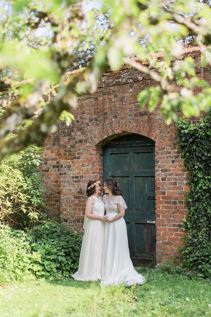 two brides standing in front of a dark green door and red brick wall in a leafy garden setting